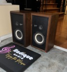 Avant 1AX speakers in walnut at an angle, grilles off, standing on tile beside the Radique board