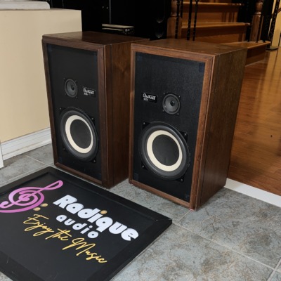 Avant 1AX speakers in walnut at an angle, grilles off, standing on tile beside the Radique board
