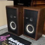 Avant 1AX speakers in walnut at an angle, grilles off, standing on tile beside the Radique board