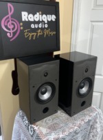 Energy Connoisseur C-2 pair seen at an angle, glossy black cabinet tops and sides beside matte front baffles with the grilles off