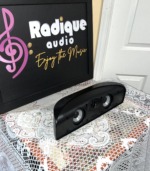 Polk Audio TL1 centre speaker in gloss black at an angle, grille off, on lace by the Radique board