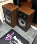 Realistic Optimus-21 speakers at an angle with grilles off, showing the walnut-grain cabinet sides and black baffles