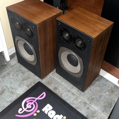 Realistic Optimus-21 speakers at an angle with grilles off, showing the walnut-grain cabinet sides and black baffles
