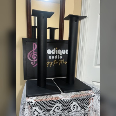 Pair of black 23.5-inch metal speaker stands with square bases and top plates, in front of the Radique Audio sign