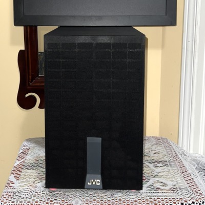 JVC SX-PW33 subwoofer standing upright, its tall narrow grille fitted with the JVC badge at the foot.