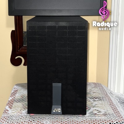 JVC SX-PW33 subwoofer standing upright, its tall narrow grille fitted with the JVC badge at the foot.