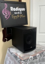 PSB Alpha SubZero i subwoofer at an angle with the metal grille on, showing the matte black cabinet side and the front control knobs