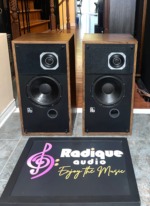 PSB Avante speaker pair with grilles off, wood veneer cabinets, square-plated tweeters, black woofers and a round port under each, on a tiled floor