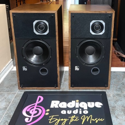 PSB Avante speaker pair with grilles off, wood veneer cabinets, square-plated tweeters, black woofers and a round port under each, on a tiled floor