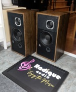 PSB Avante speakers angled with grilles removed, showing the wood veneer side panels, tweeter and woofer layout and the PSB badge on each baffle