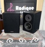 Paradigm ADP-150 surround speakers side by side, one with its black cloth grille fitted and one bare showing the tweeter and woofer