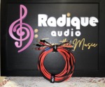 Radique House CCA speaker cable pair coiled upright in front of the Radique Audio board, red and black runs gathered by a Radique strap