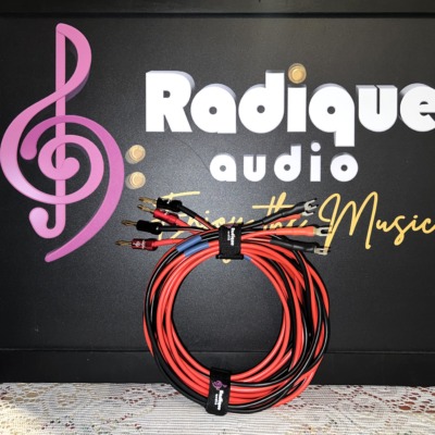 Radique House CCA speaker cable pair coiled upright in front of the Radique Audio board, red and black runs gathered by a Radique strap