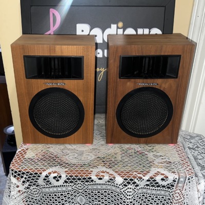 Realistic Minimus-5 speaker pair head-on, wood-grain cabinets with a black horn tweeter above a round perforated woofer grille and Realistic script badge