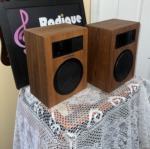 Realistic Minimus-5 speakers at an angle on a lace-covered table, showing the wood-grain side panels, horn tweeters and round woofer grilles