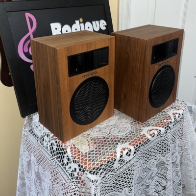 Realistic Minimus-5 speakers at an angle on a lace-covered table, showing the wood-grain side panels, horn tweeters and round woofer grilles