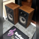Realistic Optimus-21 speakers at an angle with grilles off, showing the walnut-grain cabinet sides and black baffles
