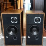 PSB Avante speaker pair with grilles off, wood veneer cabinets, square-plated tweeters, black woofers and a round port under each, on a tiled floor