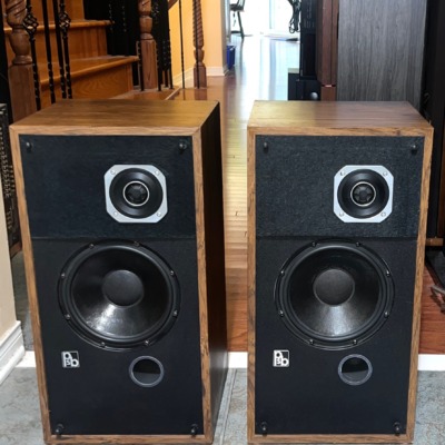 PSB Avante speaker pair with grilles off, wood veneer cabinets, square-plated tweeters, black woofers and a round port under each, on a tiled floor